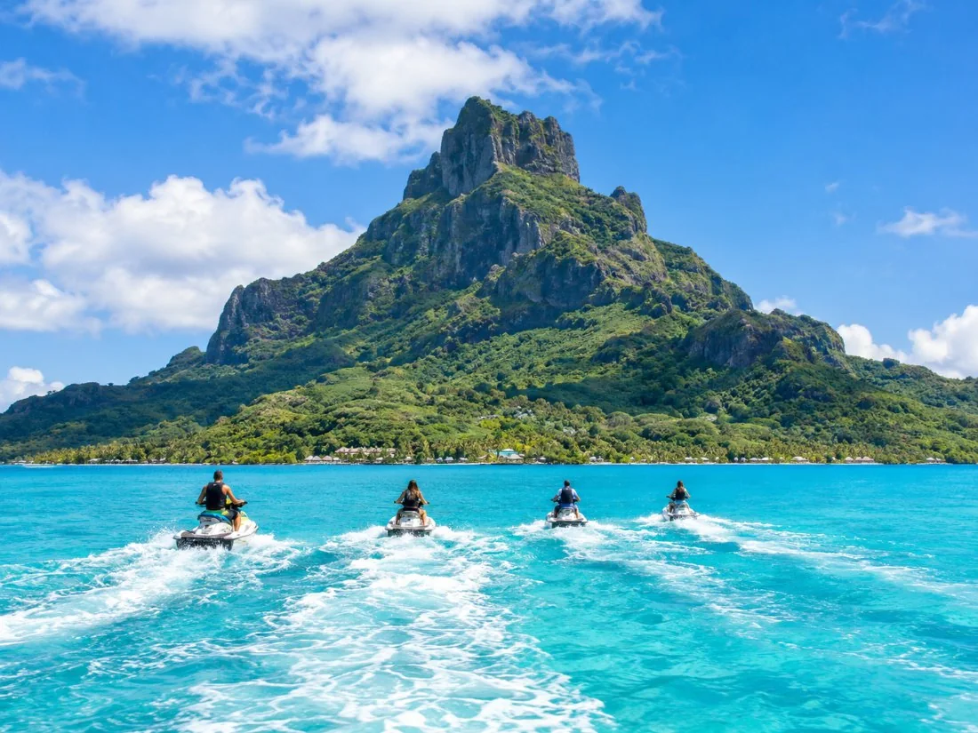 Jet Skis fahren in einer Reihe über die türkisfarbene Lagune, im Hintergrund die dunkelgrüne Spitze des Mount Otemanu